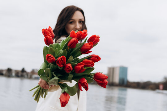 Attractive brunette woman walking on the beach shore in moody cloudy windy weather with bouquet of red tulips flowers, dressed in white suit jacket. International Women`s Day 8th March concept