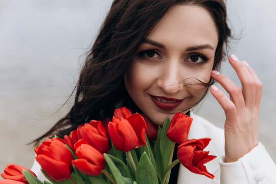 Attractive brunette woman walking on the beach shore in moody cloudy windy weather with bouquet of red tulips flowers, dressed in white suit jacket. International Women`s Day 8th March concept
