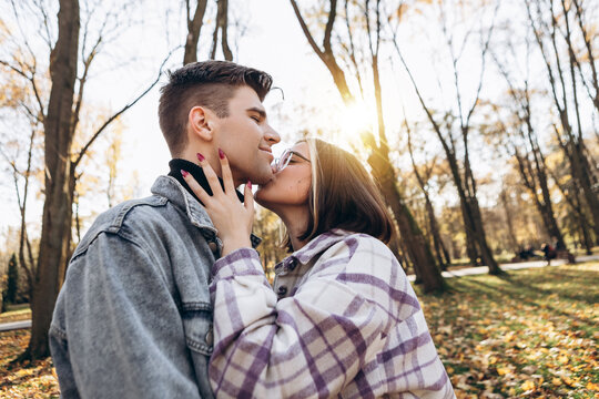 Heterosexual caucasian young loving couple walking outside in the city park in sunny weather, hugging smiling kissing laughing spending time together. Autumn, fall season, orange yellow red leaves
