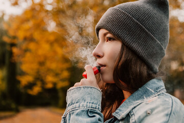 Young woman outside autumn park fall and smoking tobacco device electronic cigarette heater. Smoke and steam system with sticks inside, image with copy space. Harmful habit harm to health lungs - obrazy, fototapety, plakaty