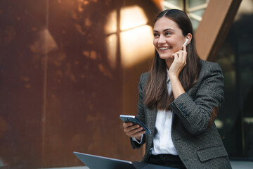 Young woman freelancer sitting outdoors with laptop and wireless headphones calling on laptop, talk by webcam, video conference. Remote work lifestyle concept