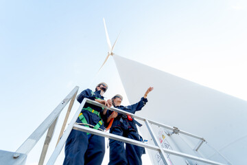 Lower view of professional technician man and woman stay on base of windmill or wind turbine and woman also point to the right with blue sky.