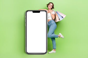 Full size portrait of attractive cheerful girl hold mall bags empty space big smart phone screen isolated on green color background