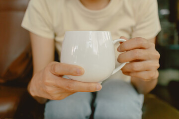 Close up of woman two hands holding white coffee cup, showing milk tea glass while sitting on brown sofa at home.