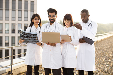 Medical specialist sharing with experience and knowledge with partners. Young adult arabian doctor who using laptop showing results of patient's tests to multiracial colleagues outdoors near clinic.