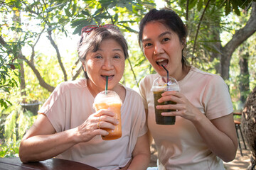 Happy asian Thai Chinese elder mother and daughter drinking cold beverages together and sitting at cafe in garden, spending good time looking at camera.