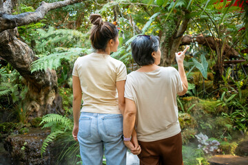 Back side of daughter and elder mother standing and enjoying time with natural forest garden together, pointing something, traveling on holidays.