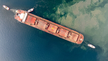 Aerial top down view of Tug boats assisting big cargo ship. Large cargo ship enters the port...