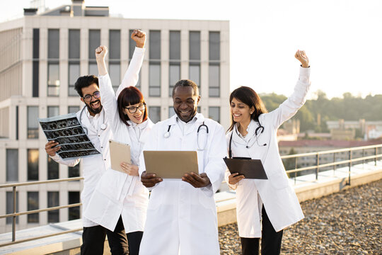 Multiethnic Successful Medical Workers Standing Outdoors Behind Young African Colleague Using Laptop And Looking Together At Results Of Patient Tests With Raising Hands. Concept Of Good Treatment.