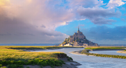 the Mont Saint-Michel  at sunset with grass and water in the foreground and cloudy sky,Normandy,...