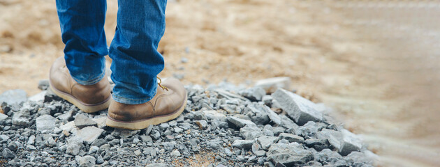 Legs of Lonely man wearing jeans and leather boots walking along the path strewn with rocks. Travel Concept-banner image. - obrazy, fototapety, plakaty