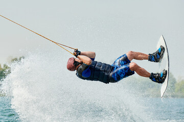 a handsome man on a wakeboard performs a backroll trick from a springboard on water with splashes