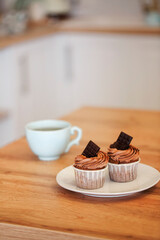 Two muffins on white plate decorated with cocoa cream and pieces of chocolate with cup of tea