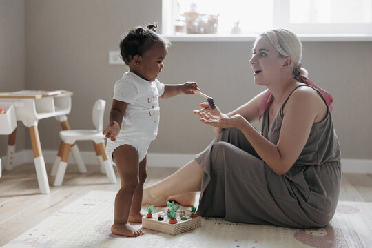 Happy caucasian mother sits in nursery and plays with her cute mixed race toddler daughter.