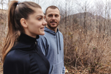 Portrait of a happy fit young couple standing in nature, wearing sportswear, ready for a run. Young female and male athlete out for a workout in pine forest.