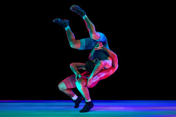 Two young men in blue and red wrestling tights wrestling and making suplex fight against black background in mixed neon lights. Concept of motion, action, combat sports, strength and power, movement.