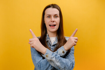 Portrait of pleasantly surprised young woman standing with crossed hands and pointing to copy space on both sides, wearing denim jacket, posing isolated over yellow color background wall in studio