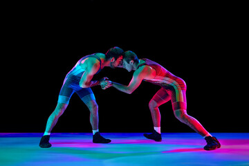 Young athlete man, wrestlers in blue and red uniform hand wrestling in neutral position on their feet in neon lights against black background. Concept of fair wrestling, championship, win competition. - obrazy, fototapety, plakaty