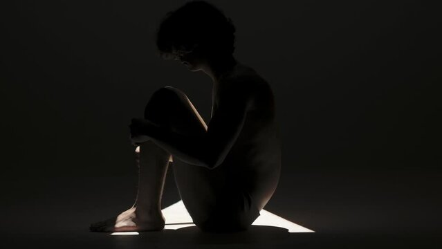 Portrait of male model silhouette on the black background in spotlight. Handsome man in underwear sits on the floor side view at camera.