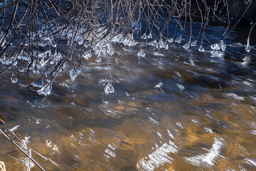 Icicles hang on branches lowered into the river. The river flows very fast. Long exposure photo of water. Winter or spring landscape
