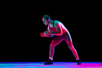 Freestyle wrestler in red fighting tights stands in attack position and looking ta camera against black background in mixed neon lights. Concept of professional sport, championship, strength, power.