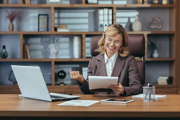 Smiling senior businesswoman in a well-appointed home office, confidently managing remote work with digital tablet and laptop.