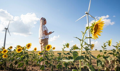 A young woman with a tablet in a field with sunflowers, wind turbines for green energy production,...