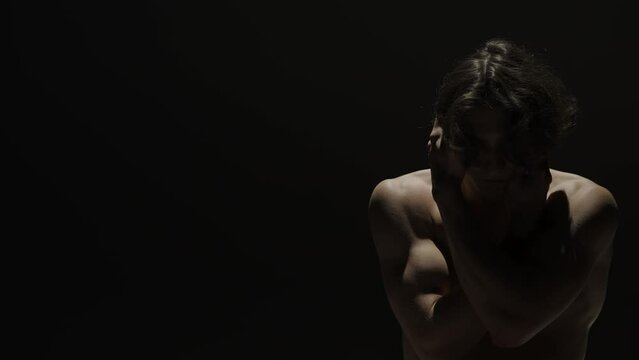 Portrait of male model silhouette on the black background in spotlight. Handsome man in underwear holds hands on face posing at camera.