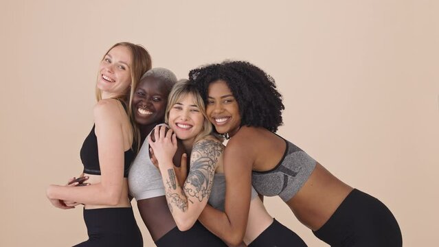 Happy fit sporty diverse young women wearing sportswear hugging standing at beige background. Multiracial girls friends advertise sport fitness gym trainings together embracing posing for portrait.