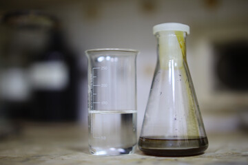 A measuring cup with a clear liquid and a flat-bottomed flask with an unknown substance standing on a dirty surface