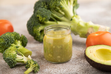 Baby puree with vegetable mix, broccoli, avocado in glass jar on brown concrete, side view,...