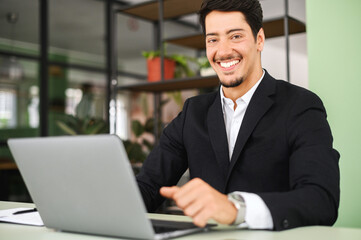 Fototapeta na wymiar Cheerful clever male latin white collar worker using laptop indoors, looking at the camera and smiles, portrait of proud and happy male office employee on the workplace
