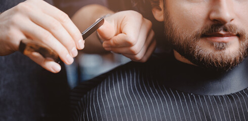 Hipster man sitting in armchair barber shop, hairdresser shaves men beard with dangerous razor