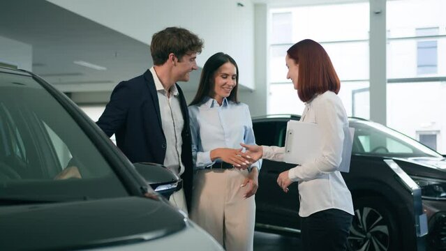 Caucasian couple clients buyers conclude agreement deal buy buying new car auto automobile in showroom rental service rent transport shaking hands with saleswoman woman dealer seller trader handshake