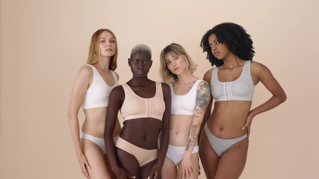 Four diverse girls hugging isolated on beige background. Group portrait. Confident multicultural young women, four multiethnic inclusive beauty models wearing underwear bonding looking at camera.