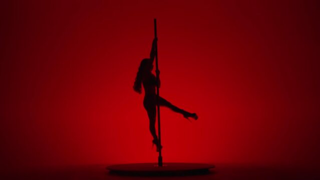 Dark silhouette of female dancer performing pole dance in studio on red background. Woman in high heeled shoes performs rotation on a pole.