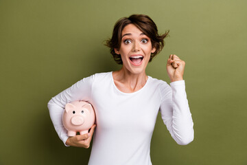 Photo portrait of pretty young girl hold piggy account raise fist winning dressed stylish white outfit isolated on khaki color background