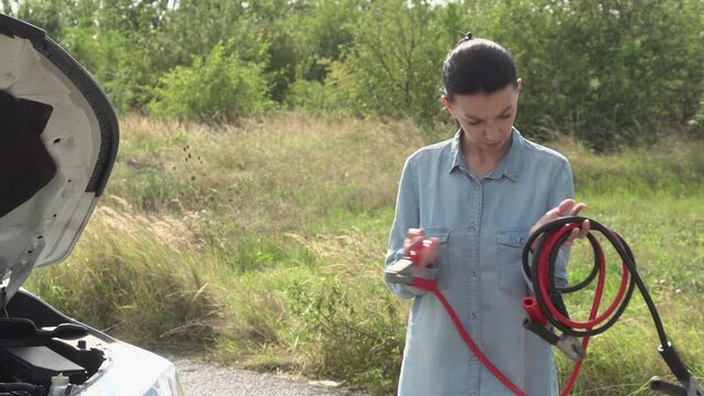 A female driver stands by the roadside next to a broken-down car, attempting unsuccessfully to jumpstart the vehicle by connecting ignition cables.