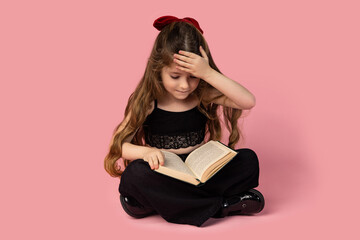 The amazed girl sitting with her legs crossed and her hand on her forehead, dressed in black, studying a book, isolated on a pink background.