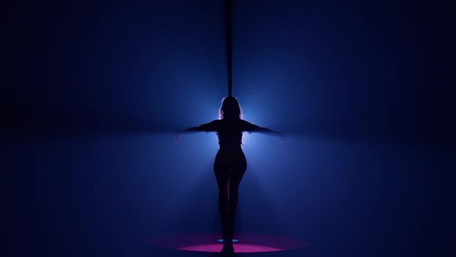 Dark silhouette of a flexible and plastic woman dancing on a pole. Woman dancing on a pylon in a dark studio against a bright spotlight.