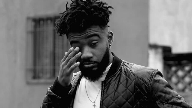 One regretful young black man covers face with hand while standing outside in street. African American person rubbing eyes in black and white, monochromatic