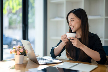 Freelance Asian woman enjoying morning coffee during break from work with sunlight shining through...
