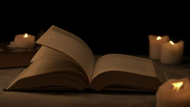 Close shot of retro hardcover book on dark background in warm light. Air blows dusty pages of vintage book surrounded by candles.