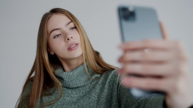 Confident attractive woman in warm sweater hold mobile phone in hand and make selfie on white background. Young beautiful female posing and take self picture on smartphone.