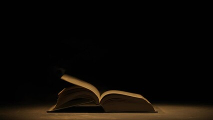 Close up shot of retro hardcover book on dark background in warm light. Vintage book laying on the ground wind blowing off pages.
