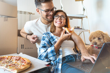 Cheerful young couple wearing eyeglasses while working on laptop and eating pizza with dog sitting...