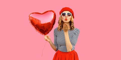 Portrait of happy young woman with red heart shaped balloon blowing her lips wearing french beret on pink studio background