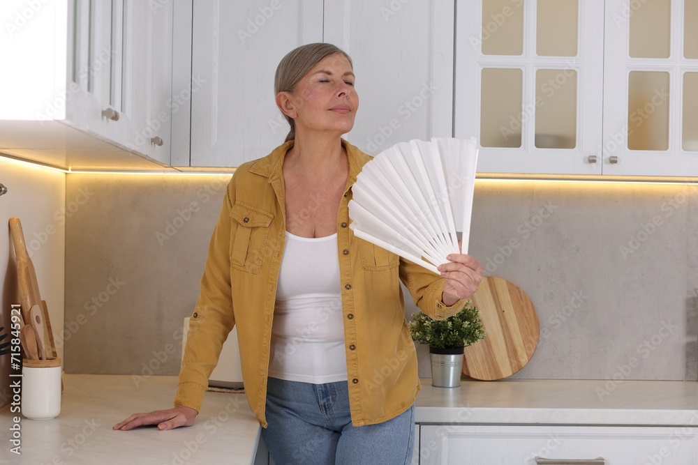 Poster Menopause. Woman waving hand fan to cool herself during hot flash in kitchen