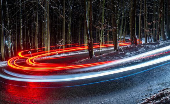 Country road at night with white and red light traces of cars driving through darkness. Long time exposure of a winding forest road with hairpin curve, reflecting wet street and snowy beech trees.