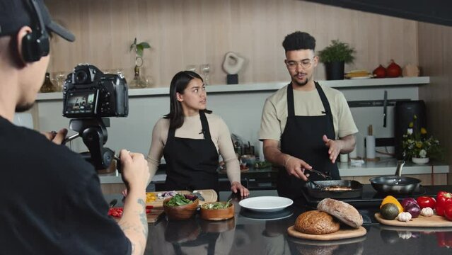 Young male chef frying meat steak and explaining recipe to female co-host while cameraman shooting cooking TV show in kitchen